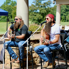 Two musicians playing guitar on stools with microphones in an outdoor setting.