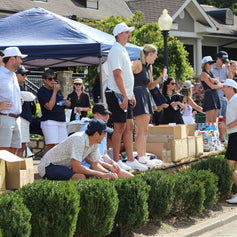 People gathered around a table with boxes at an outdoor event, under blue tents.